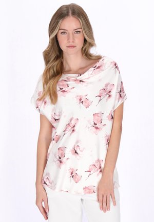 Tânără cu păr lung, ondulat și blond, purtând o bluză albă cu modele florale roz și pantaloni albi, pe un fundal simplu.