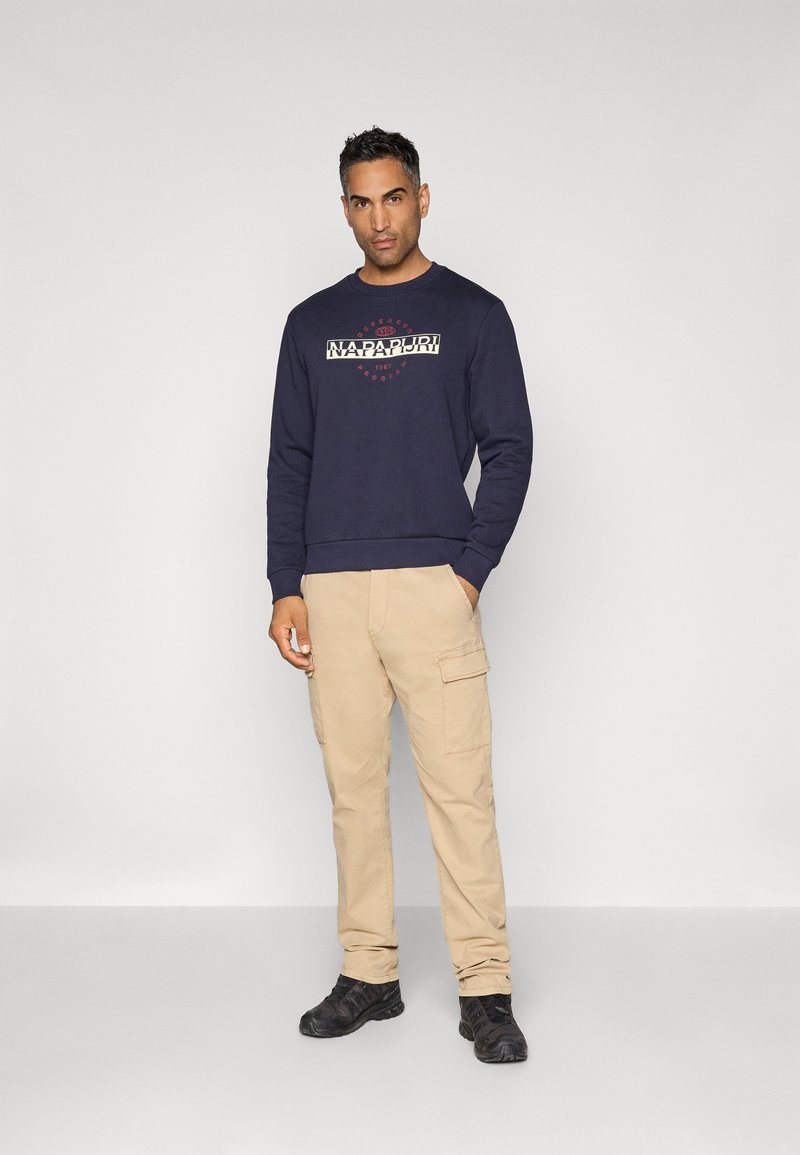 Un homme portant un sweat à capuche bleu marine avec le logo "Napapijri", un pantalon cargo beige et des baskets noires, se tenant contre un fond gris clair uni.