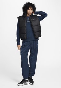 Juodas pūkuotas liemėlis užsisenėjusio mėlyno megztinio fone, derinamas su mėlynomis kelnėmis. Ypatybės apima užtrauktuką ir Nike prekės ženklą. Apsivilkta su juodais sportbačiais.