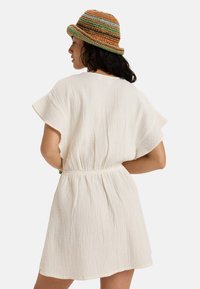 White, crinkled fabric dress with wide sleeves and a cinched waist. A multicolored woven hat complements the outfit.