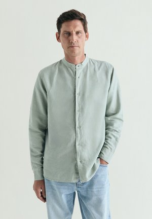 Hombre con camisa verde claro abotonada con cuello mandarín y vaqueros azul claro, de pie con una mano en el bolsillo delante de un fondo liso.