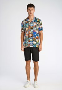 Korte mouwen, multikleurig shirt met verschillende reisgerelateerde prints. Gecombineerd met zwarte shorts en witte sneakers.