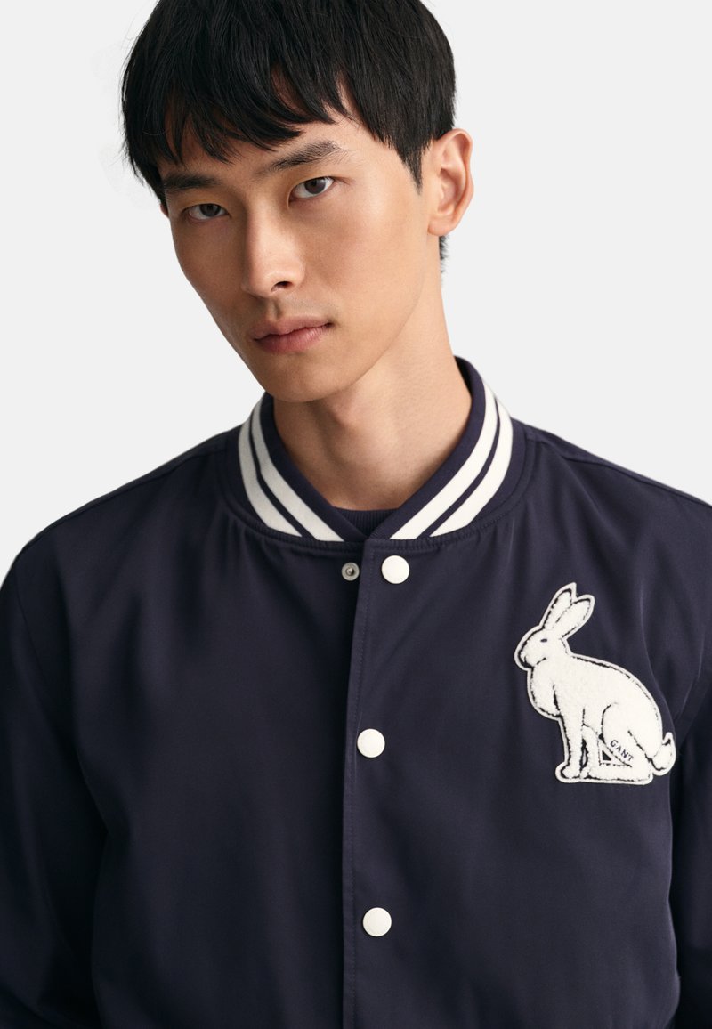 Marineblauer Bomberjacke mit einem kontrastierenden gestreiften Kragen, versehen mit einem weißen gestickten Hasen auf der linken Brust und einem Druckknopfverschluss.