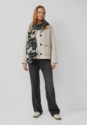 Donna che indossa una giacca beige chiaro, una sciarpa a fantasia, un maglione crema, jeans neri a gamba larga e sneakers bianche, in piedi davanti a uno sfondo grigio uniforme.