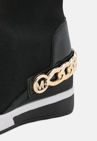 Baskets compensées noires avec semelle blanche, détail chaîne dorée et logo circulaire doré avec les initiales "MK" près du talon.