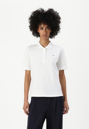 Žena s prirodno kovrčavom kosom nosi bijelu polo majicu kratkih rukava i tamne nabrane hlače, stoji ispred jednostavne bijele pozadine.