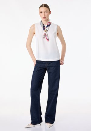 Kvinde iført en ærmeløs hvid top, mørkeblå vidde jeans, hvide spidse hæle og et rødt, hvidt og blåt stribet halstørklæde med gulddetalje.