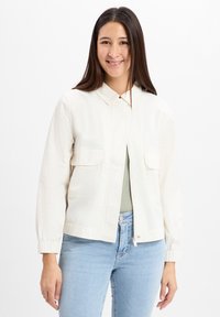 Lichtgekleurde crèmekleurige zip-up jas met kraag, twee voorzakken en elastische boorden, gecombineerd met lichtblauwe jeans. Glad, katoenen materiaal.