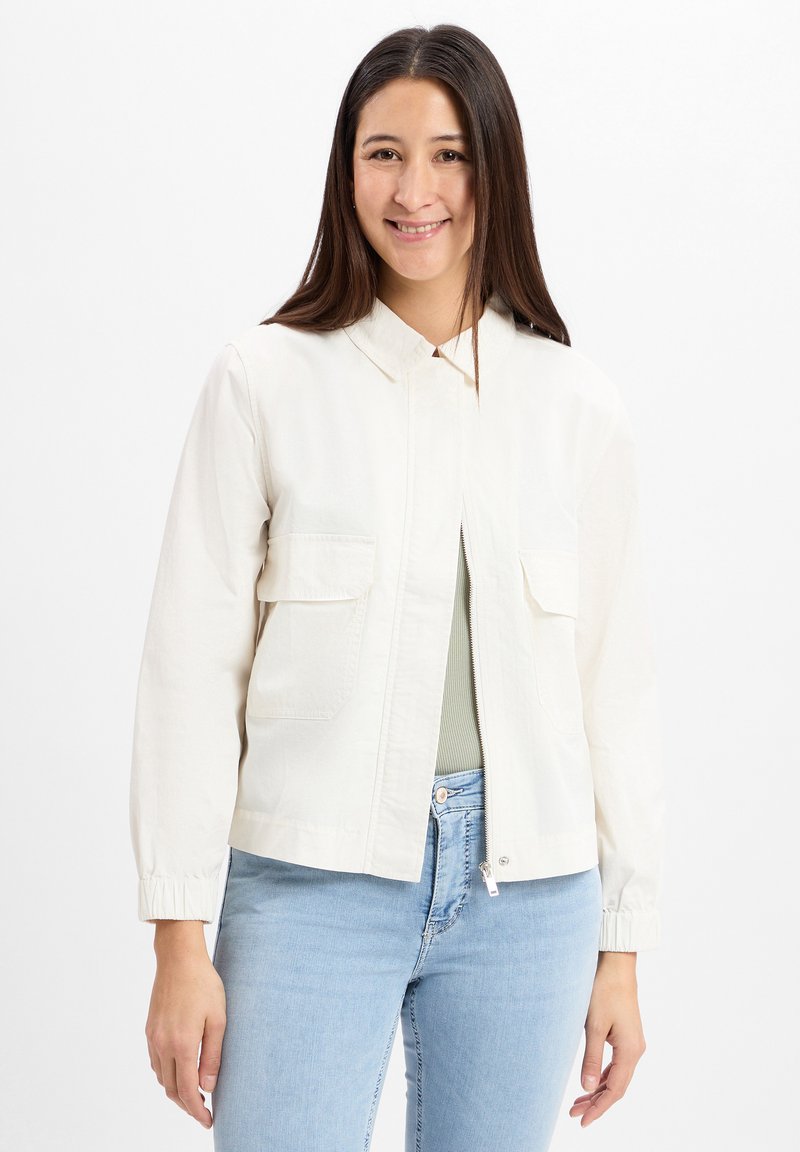 Lichtgekleurde crèmekleurige zip-up jas met kraag, twee voorzakken en elastische boorden, gecombineerd met lichtblauwe jeans. Glad, katoenen materiaal.