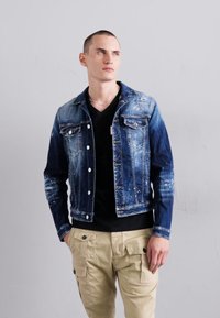 Chaqueta de mezclilla con un degradado de claro a oscuro, textura desgastada y herrajes plateados. Llevada sobre una camiseta negra y pantalones cargo beige.