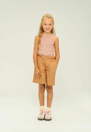 Tan shorts met zijzakken, gecombineerd met een roze, mouwloos top met een bloemenpatroon. Draagt lichte sokken en twee-kleurige sneakers.