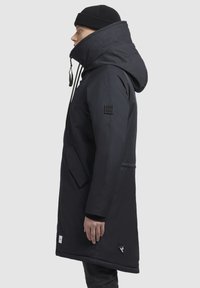 Abrigo largo negro con cuello alto, cremallera frontal, capucha con cordón ajustable y bolsillos laterales. Presenta un parche con el logotipo en la manga y el dobladillo.