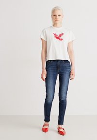 Weißes Baumwoll-Cropped-T-Shirt mit rotem Vogel-Graphic, kombiniert mit dunkelblauen Skinny-Jeans und roten Ballerinas. Sanfte Texturen überall.