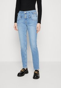 Vaqueros de mezclilla azul claro con cintura alta y cierre de tres botones, que cuentan con bolsillos delanteros y un diseño ajustado. Combinados con mocasines negros.