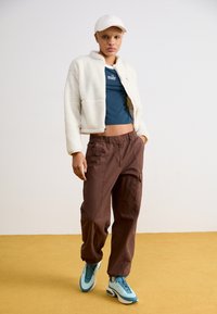 Weiße Fleece-gekürzte Jacke über einem dunkelblauen Oberteil, braune weit geschnittene Hose und blaue Turnschuhe, vor einem hellen Hintergrund und einem sandfarbenen Boden.