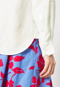 Camicia di lino bianca con orlo curvo e polsini abbottonati, abbinata a una gonna blu con un audace motivo floreale rosso.