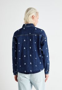 Casaco de ganga azul escuro com bordados florais brancos. Apresenta um colarinho, punhos com botão e bainhas cosidas. Corte descontraído com uma pala nas costas.