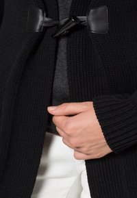 Czarny dziergany kardigan o prążkowanej teksturze, z zapięciem na skórzany zamek. Wewnętrzna warstwa jest szara, ma luźny krój i otwarty przód.