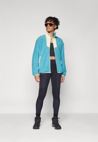 Veste en polaire teal avec un col crème, fermeture éclair à l'avant, portée avec des leggings noirs et des bottes. Modèle portant des lunettes de soleil oversize.