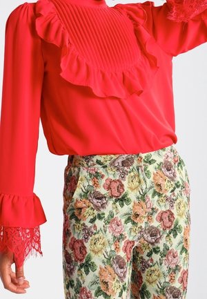Blusa rossa a maniche lunghe con rouches e polsini in pizzo abbinata a pantaloni a vita alta in arazzo floreale nei toni del rosa, giallo e verde.