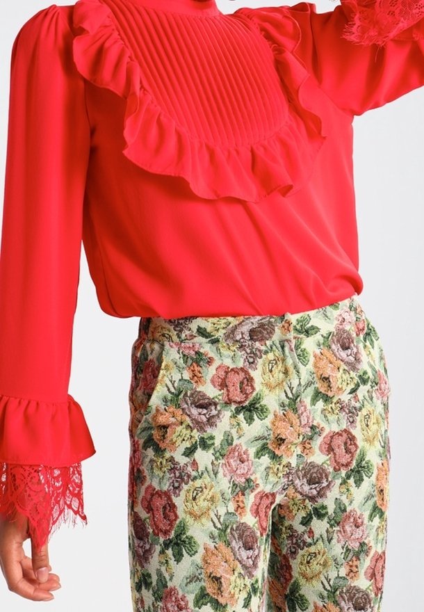 Blusa rossa a maniche lunghe con rouches e polsini in pizzo abbinata a pantaloni a vita alta in arazzo floreale nei toni del rosa, giallo e verde.
