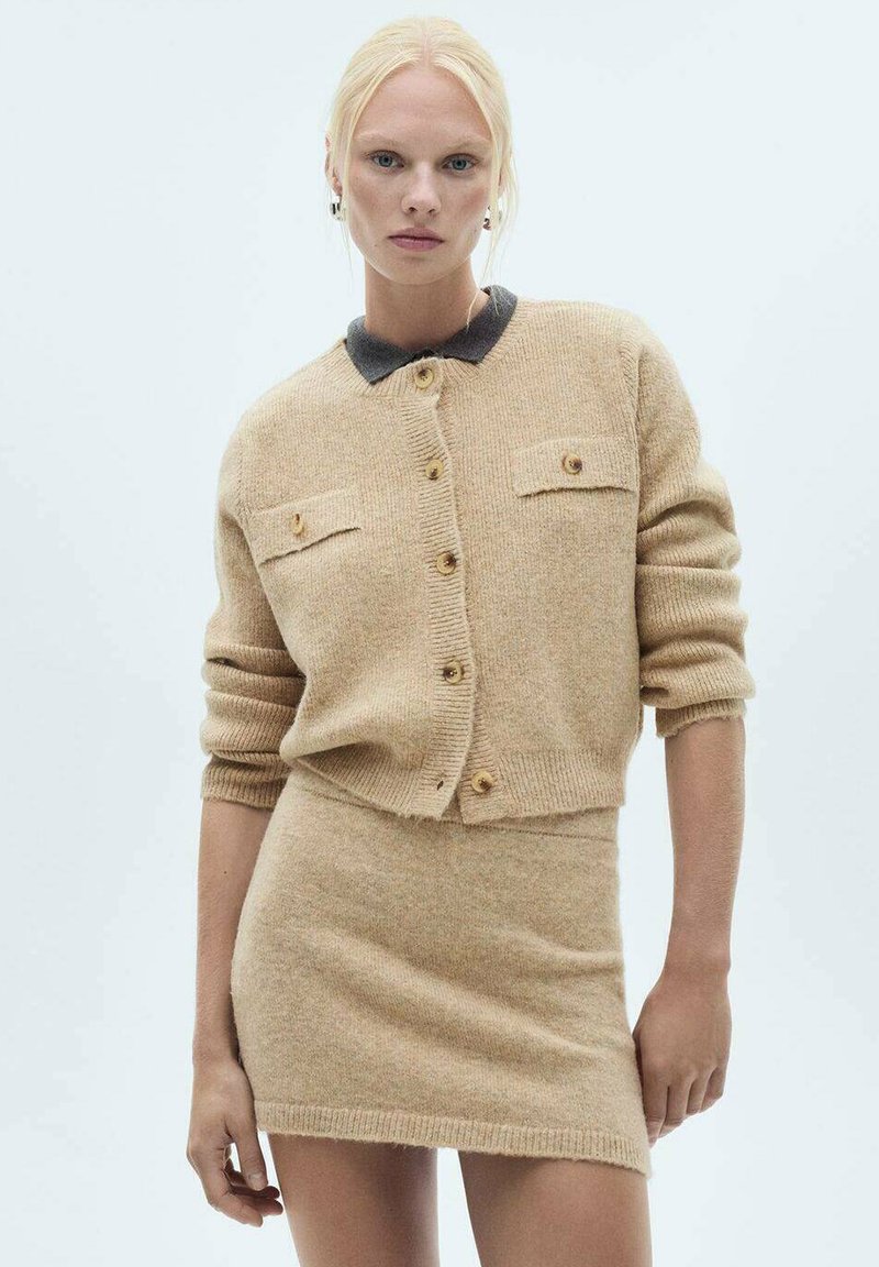Beige stickad kofta med krage, knappstängning och två bröstfickor, tillsammans med en matchande figursydd mini kjol. Slät textur, mjukt tyg.