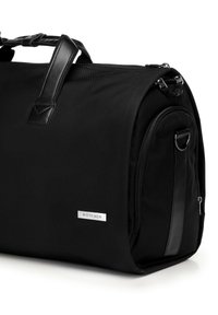 Schwarze Sporttasche aus strapazierfähigem Gewebe mit Lederakzenten, einem Reißverschluss und einem flachen Boden. Mit einer markierten Metall-Logo-Platte.