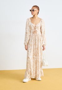Robe maxi en beige avec un motif floral, dotée d'un décolleté en V, de manches longues et d'une jupe froncée. Portée avec des baskets blanches et des lunettes de soleil.