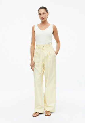 Frau trägt ärmelloses weißes Top, beige weite hoch taillierte Hose mit Gürtel und Sandalen, steht mit einer Hand in der Tasche.