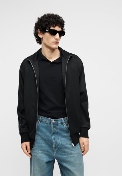 Άνδρας που φοράει μαύρο μπουφάν με φερμουάρ, μαύρο πουκάμισο, μπλε τζιν και ορθογώνια μαύρα γυαλιά ηλίου, στέκεται μπροστά σε ένα απλό φωτεινό φόντο.