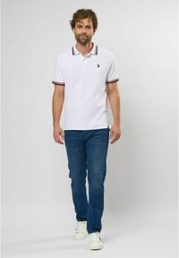 Hvid polo skjorte med kontrasterende sort og rød stribet krave, parret med blå jeans og hvide sneakers. Enkelt, afslappet design.
