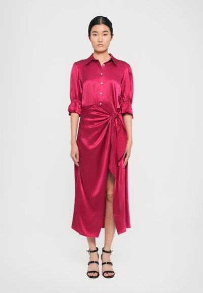 Femme portant une robe midi en satin magenta avec des manches bouffantes trois-quarts, des boutons sur le devant, un nœud sur le côté et des talons strappy noirs.