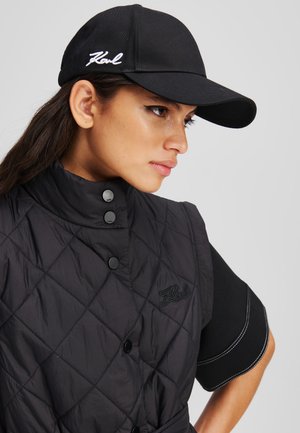 Schwarze gep quiltete Weste mit hohem Kragen und schwarzen Knöpfen, kombiniert mit einer schwarzen Mütze mit weißem, gesticktem Logo. Glattes, strukturiertes Material.