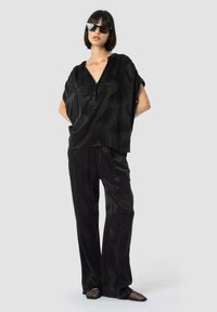 Črno, ohlapno oblačilo z zgornjim delom na gumb, ki ima subtilen vzorec, in širokimi hlačami; dopolnjeno z črnimi, kotnimi sončnimi očali.