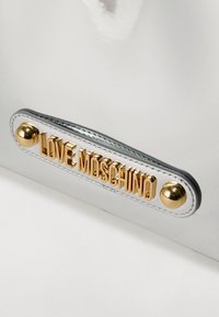 Τσάντα χειρός από ασημένιο πατέν δέρμα με πλακέτα λογότυπου σε χρυσαφί απόχρωση και στρογγυλές μεταλλικές διακοσμήσεις. Ομαλή υφή με ραφές για λεπτομέρεια.