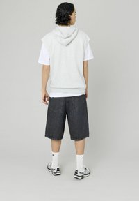 Haut à capuche sans manches gris clair sur un t-shirt blanc, associé à un short en denim foncé. Modèle portant des chaussettes blanches et des baskets noir et blanc.