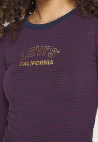 Nahaufnahme einer Person, die ein gestreiftes Levi’s California Hemd in Marineblau und Burgunderrot sowie eine zarte goldene Perlenkette trägt.
