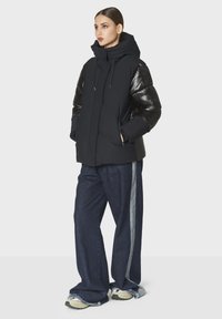Giacca a vento nera con maniche lucide, cappuccio regolabile e tasche laterali, abbinata a pantaloni in jeans scuri a gamba larga con una striscia laterale.