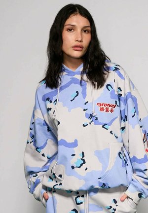 Mujer joven con cabello largo y oscuro, vistiendo una sudadera con capucha suelta de camuflaje azul y blanco con texto rojo, manos en los bolsillos, fondo neutro.