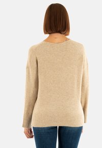Mujer con pelo castaño liso llevando un suéter de manga larga beige y jeans azules, de pie de espaldas contra un fondo blanco.