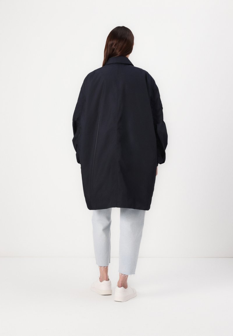 Abrigo negro de gran tamaño con un cuello estructurado, mangas largas, tejido suave y un ajuste relajado, acompañado de jeans cortos de color azul claro y zapatillas blancas.