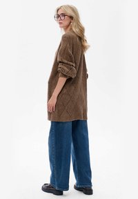 Kastanjebruine gebreide oversized trui met een ingewikkeld diamanten patroon, gecombineerd met wijde blauwe jeans en forse zwarte schoenen.