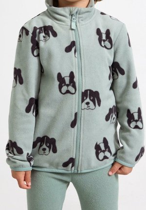 Dziecko ubrane w jasnorozpinaną zieloną polarową kurtkę i spodnie, kurtka w wzór z czarno-białymi twarzami psów, stojące na jednolitym tle.