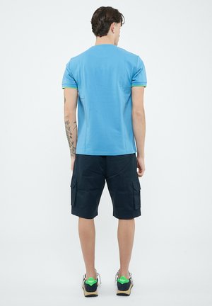 Uomo di spalle che indossa una maglietta blu, pantaloncini cargo neri, scarpe da ginnastica, con un tatuaggio floreale sull'avambraccio sinistro, in piedi su uno sfondo semplice.