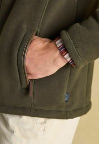 Olivovo zelená fleecová bunda so zipsovým vreckom a textúrovanou látkou. Na zápästí je viditeľná manžetová košeľa s multikolórovou károvanou vzorom.