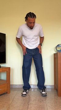 Jeans de ganga azuis com um corte reto, combinados com uma t-shirt branca de manga curta e ténis pretos, de pé sobre um chão de azulejos.