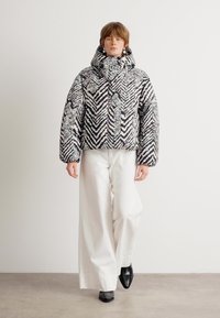 Giacca a vento con motivo zigzag in bianco e nero, caratterizzata da un collo alto e maniche a palloncino, abbinata a pantaloni larghi bianchi e scarpe nere.