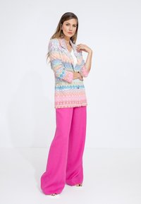 Blazer lavorato a maglia multicolore con un motivo geometrico, abbinato a pantaloni larghi rosa acceso e scarpe bianche, su uno sfondo neutro.