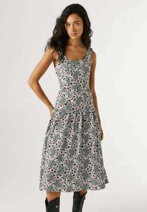 Frau trägt ein ärmelloses, eng anliegendes Kleid mit floralem Muster in Grün- und Brauntönen, kombiniert mit schwarzen Stiefeln bis zur Mitte der Wade, steht vor einem einfarbigen Hintergrund.