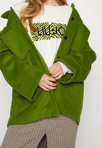 Cappotto oversize in misto lana verde con grandi tasche e chiusura a bottoni, indossato sopra un maglione bianco con motivo a zebra.